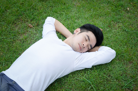 Portrait of a young happy Asian man relaxing on the grass with his hands under the headの写真素材