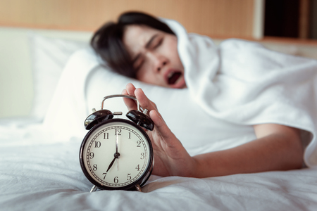 Turning off Sound or Snoozing The Alarm Clock Concept, Beautiful Woman Waking Up in The Morning at Bedroom While Her Hand is extending Alarm of Timer Clock.の写真素材