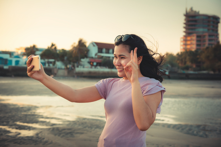Traveling and Technology Concept, Woman is Taking Selfie Photos at The Beach by Her Smartphone in Summer Vacation. Asian Tourist is Relaxing With Her Cell Phone on The Beach in Holiday Time.の写真素材