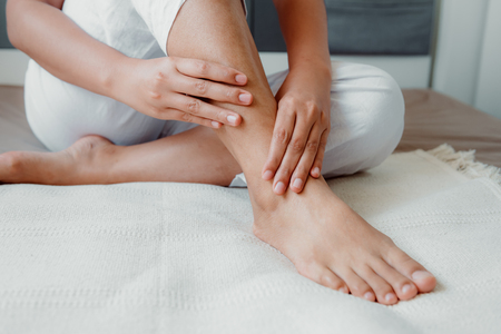 Close-Up of Woman Hands is Massaging Her Ankle on Bedroom, Young Adult having Legs Ache After Home Work. Healthcare and Medicine Concept.の写真素材