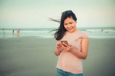 Portrait of Woman is Using Smartphone at The Beach During Sunset, Asian Tourist is Relaxing With Her Cell Phone on The Beach in Holiday Time. Technology Communication Concept.の写真素材