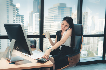 Technology Communication Concept, Portrait of Attractive Business Woman is Calling on Cellphone While Working on Her Table Desktop in Front of Windows Office Building Overlooking The City View.の写真素材