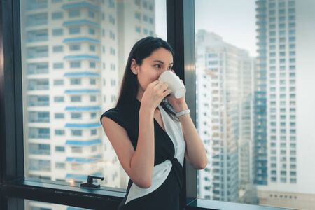 Portrait of Businesswoman is Talking to Someone on Mobile Phone and Drinking a Cup of Coffee While Standing Against a Window Overlooking The City, Business Financial and Technology Communication.の写真素材