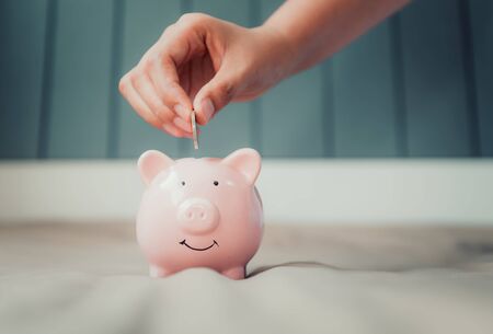 Woman Hand is Putting a Money Coin into Piggy Saving on The Bedroom. Female Hand is Inserting Coin in Piggybank. Business Banking and Financial Concept.の写真素材