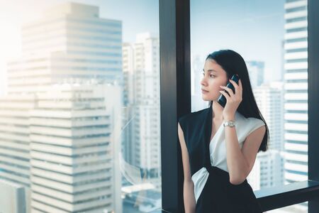 Portrait of Business Woman is Talking on Mobile Phone in Her Office., Beautiful of  Asian Woman is Calling to Someone on Her Cell Phone in Front of Transparent Windows Glass Overlooking City Downtown.の写真素材