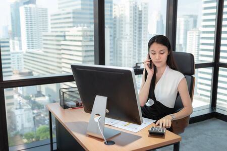 Business Woman Using Calculator and Talking on Mobile Phone in Her Office., Beautiful of  Asian Woman is Calling to Someone on Her Cell Phone While Calculating Data Saving at Her Table Desktop.の写真素材