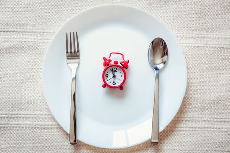 Healthy Eat Meal Time and Reminder Breakfast Concept, Food Timing Cycles for Eating, Red Alarm Clock With Stainless Steel Spoon and Fork on Ceramic Dish. Empty Plate and Household Object on a Tableの写真素材