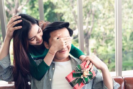 Beautiful Woman is Surprising With Gift Box While Closing Her Boyfriend Eyes at Their Home. Handsome Man Having Happy Surprise While Waiting Girlfriend Giving Presents Box. Couple Valentine Conceptの写真素材