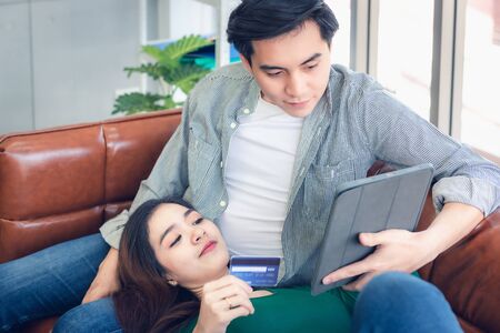 Young Couple Love Having Enjoyment While Online Shopping on Electronic Tablet in Living Room, Portrait of Asian Couple Relaxing on a Couching During Shopping Online Togetherness. Relaxation/ Lifestyleの写真素材