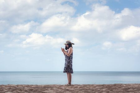 Portrait of Asian Woman is Using Mobile Phone Against Blue Sky and Sand Beach, Beautiful Woman is...