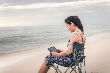 Freelancer Woman is Working on Tablet While Travel Vacation Trip at The Beach, Smart Woman is Using Tablet for Communication and Entertainment at Outdoor Sand Beach. Freelance and Relaxation Concept.の写真素材