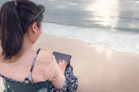 Freelancer Woman is Working on Tablet While Travel Vacation Trip at The Beach, Smart Woman is Using Tablet for Communication and Entertainment at Outdoor Sand Beach. Freelance and Relaxation Concept.の写真素材