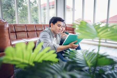 Portrait of Young Couple Love in Romantic Emotions While Reading a Book Together, Couple Young People Having Fan and Relaxing in Living Room. Happy Emotion and Relaxation Lifestyles Conceptの写真素材