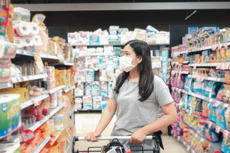 Customer Asian Woman Wearing Face Mask With Shopping Cart in Supermarket Department Store Shop While Choosing and Looking Goods on Shelf During Covid-19 Pandemic. Coronavirus Covid Situationの写真素材