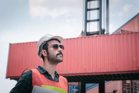 Portrait of Confident Transport Engineer Man in Safety Equipment Standing in Container Ship Yard. Transportation Engineering Management and Container Logistics Industry, Shipping Worker Occupationの写真素材