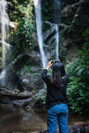 Tourist Woman is Enjoying While Photographing Waterfall on Travel Vacation, Rear View of Asian Woman Relaxing Outdoor Adventure Tourism and Leisure Activity Lifestyle. Holiday Outdoors Tripの写真素材