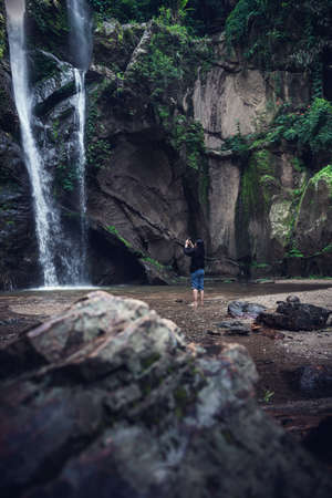 Tourist Woman is Enjoying While Photographing Waterfall on Travel Vacation, Rear View of Asian Woman Relaxing Outdoor Adventure Tourism and Leisure Activity Lifestyle. Holiday Outdoors Tripの写真素材