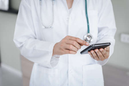 Female Doctor Using Mobile Phone for Medical Appointment Check at Hospital, Medicine Doctor Texting Message on Smartphone in Front of Examination Office. Medic Healthcare and Doctors Service.の写真素材