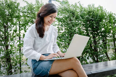 Close-Up of Woman Using Laptop for Business Communication While Sitting on Garden Bench. Businesswoman Use Computer Laptop for Remote Owner Business at The Backyard. Technology Communications Onlineの写真素材