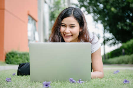 Beautiful Woman Relaxing and Using Laptop While Laying on Grass Field, Asian Woman Working Online on Laptop Device and Joying 
Social Media. Relaxation Leisure Activities Lifestyles/Work at Homeの写真素材