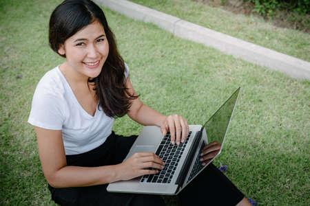 Beautiful Woman Relaxing and Using Laptop While Sitting on Grass Field, Asian Woman Working Online...