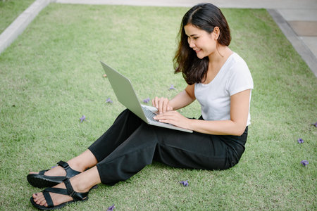 Beautiful Woman Relaxing and Using Laptop While Sitting on Grass Field, Asian Woman Working Online on Laptop Device and Joying 
Social Media. Relaxation Leisure Activities Lifestyles/Work at Homeの写真素材