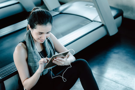 Sporty Woman in Sportswear is Sitting on Flooring While Using Tablet After Jogging Workout Break. Sport Asian Woman is Resting on The Floor and Enjoy With Mobile Tablet in Fitness Gym. Sport Lifestyleの写真素材
