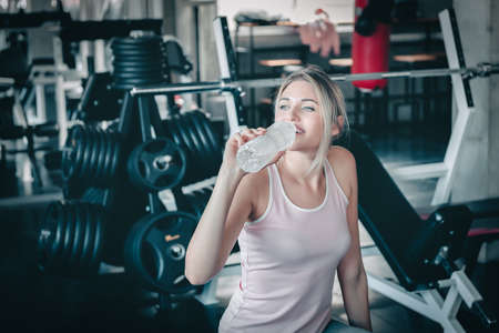 Beautiful Woman is Drinking Water From Bottle After Working Out in Fitness, Portrait of Athletic Woman is Drinking Water After Exercising in Front of Bodybuilder Machine in Gym. Sport Exercise Conceptの写真素材