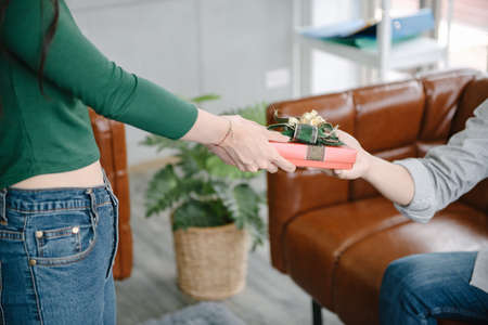 Close-Up of Couple Love Giving Present Together, Young Girl Surprise Gift to Her Boyfriend in The Living Room. Young Couple Love Give Present Box for Anniversary New Year Celebrate Together.の写真素材