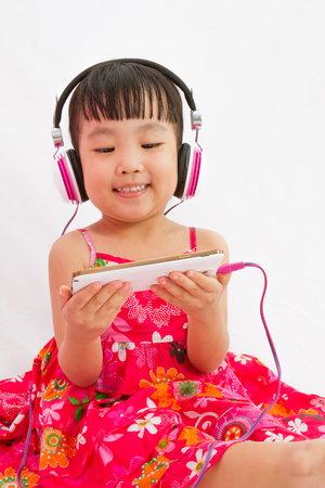 Chinese little girl on headphones holding mobile phone in plain isolated white background.の写真素材