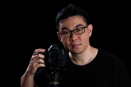 Asian Chinese Male Photographer Holding Digital SLR Camera in isolated black background.