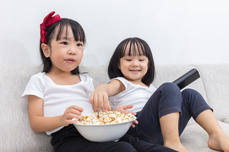 Happy Asian Chinese little sisters eating popcorn on the sofa in the living room at home.の写真素材