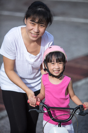 Asian Chinese little girl riding bicycle with mom guide on tar road outdoor.の写真素材