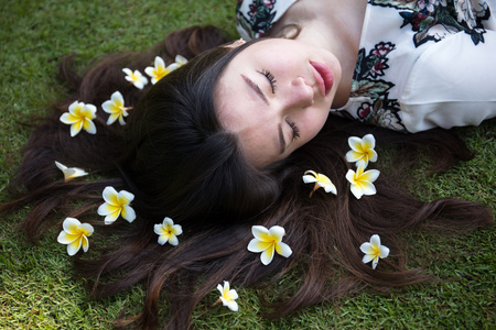 Asian beautiful young woman lying on lawn with flowers on hair in the garden on a sunny dayの写真素材