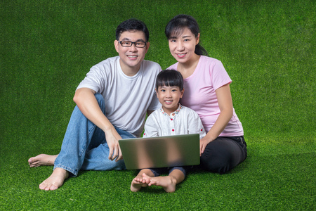 Asian Chinese parents and daughter sitting on the grass and playing laptop at outdoor parkの写真素材