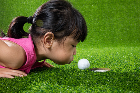 Asian Chinese little girl lying on grass and blowing the ball into a holeの写真素材