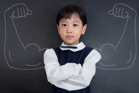 Asian Chinese little girl standing against blackboard with sketched strong and muscled arms in isolated black backgroundの写真素材