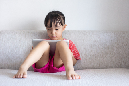 Asian Chinese little girl sitting on the sofa playing digital tablet in the living room at homeの写真素材