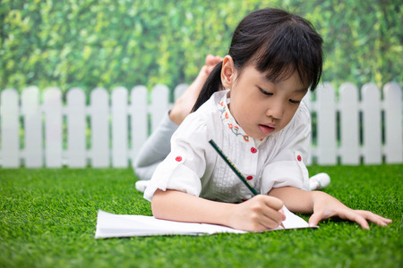 Asian Little Chinese girl kneeling on the grass and doing homework at outdoor parkの写真素材