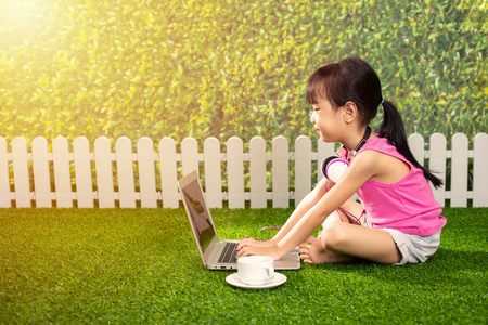 Asian Little Chinese girl sitting on the grass and playing with laptop at outdoor parkの写真素材