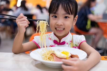 Asian Chinese little girl eating noodles with chopsticks in restaurantの写真素材
