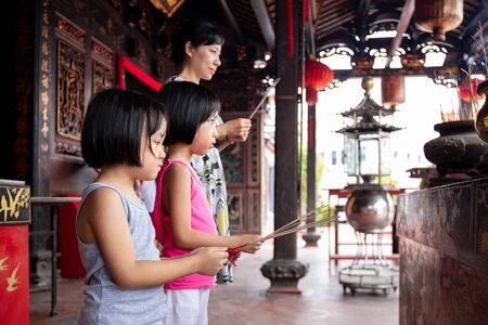 Asian Little Chinese Sisters and mother praying with burning incense sticks at a outdoor templeの写真素材