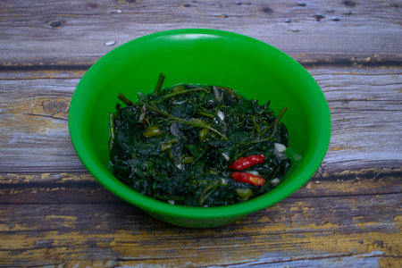 Stir-fried cassava shoots and chilies served in a green bowl on a rustic wooden background.の写真素材