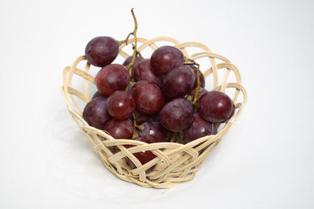 A bunch of fresh red grapes placed in a small woven basket on a white background.の写真素材