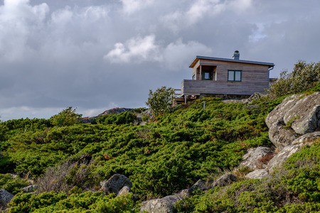 small wooden house at the top of the cliff with green bushes in the foreground and blue sky with white clouds in the backgroundの写真素材