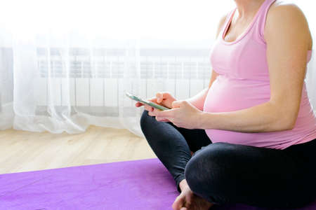 pregnant caucasian woman looking at cell phone while practicing yoga with pink t-shirt and white backgroundの写真素材