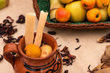 Jar of Christmas fruit punch with cane, tejocote and guava. Basket with fruits, apple. On the table tamarind and hibiscus leavesの写真素材