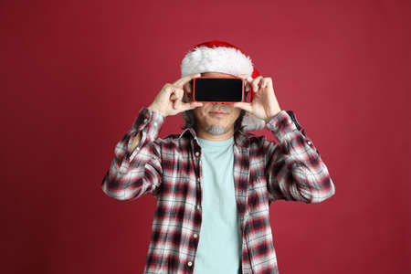 The senior Asian man wears red plaid shirt with Santa Claus hat standing on the red background.