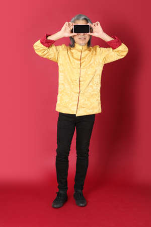 The senior Asian man wearing traditional gold chinese dresses standing on the red background.