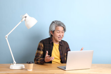 The senior Asian man wearing yellow plate shirt working on laptop at the working desk with the blue background.の写真素材